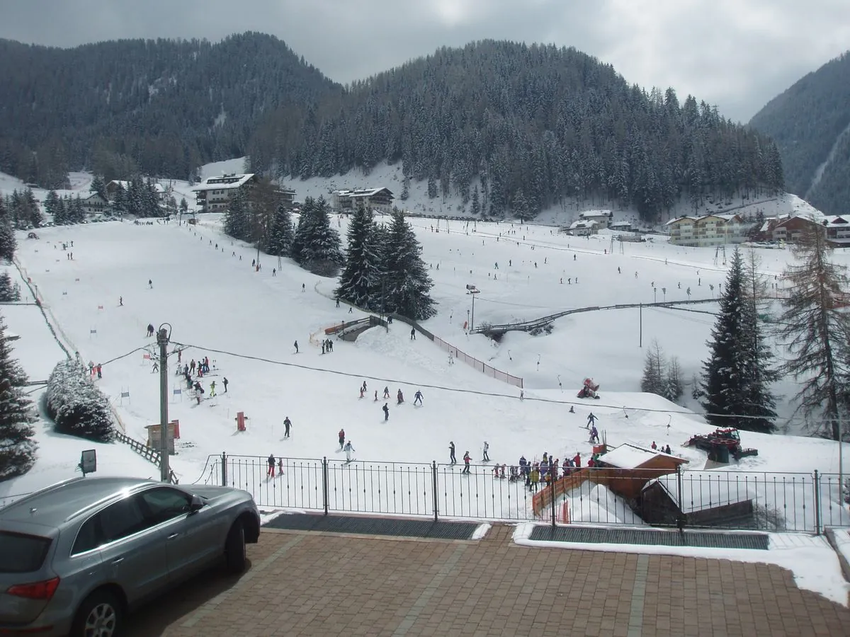 Blick auf einen Übungshang mit Skischule und Lift, vorne ein Vorplatz mit Auto