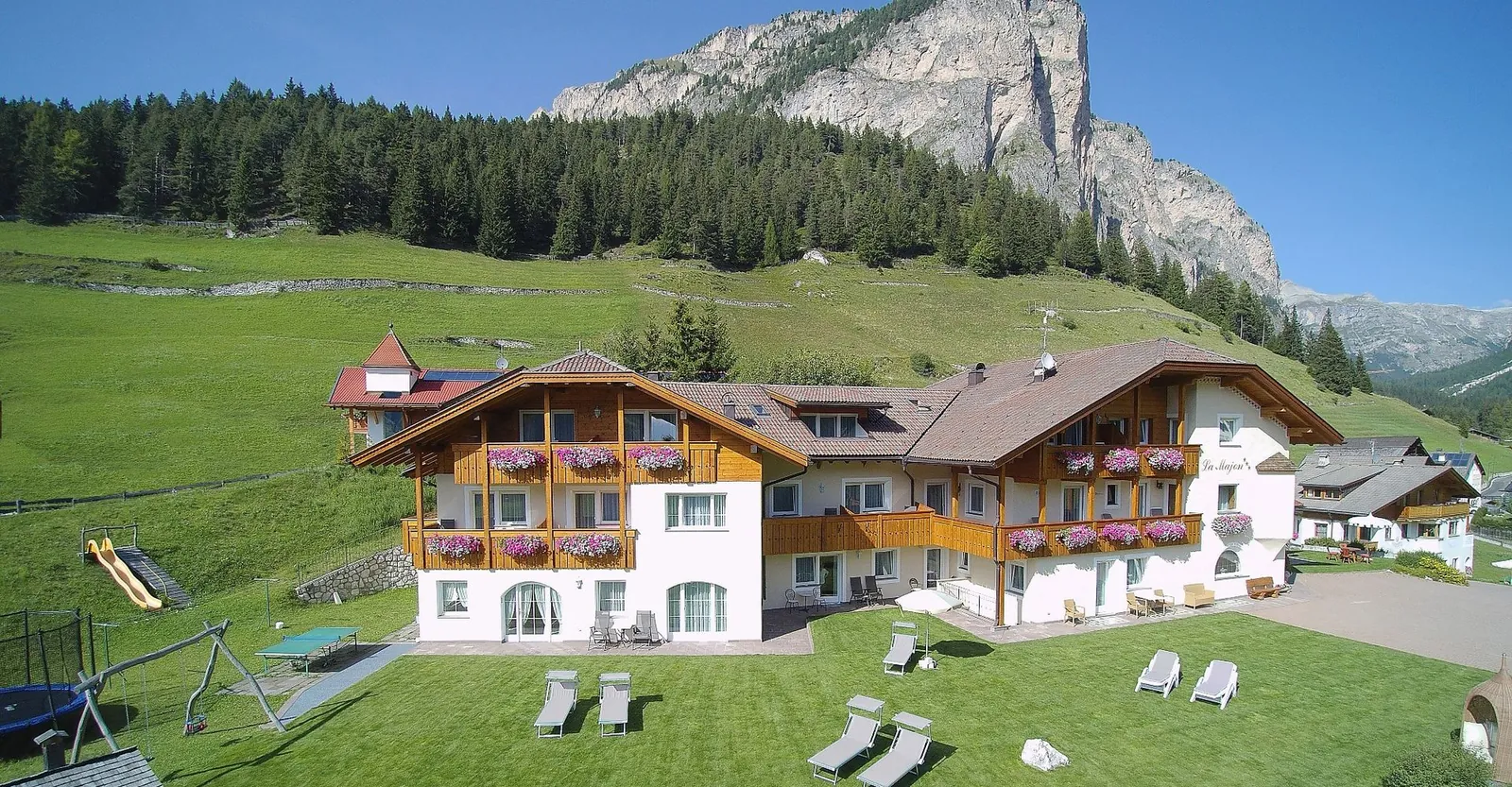 La Majon im Sommer aus der Luft, mit Liegewiese, Spielplatz und Blumenbalkonen