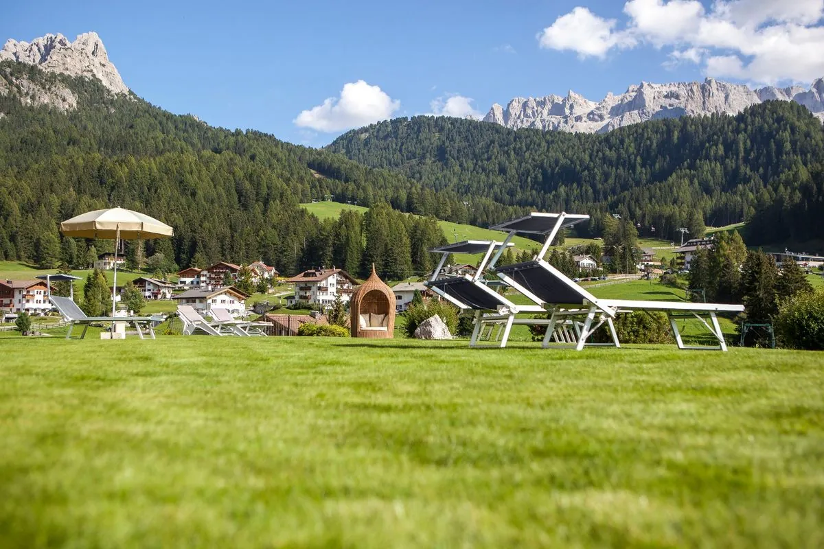 Liegewiese mit Sonnenliegen und Schirm, dahinter Wald und Dolomitengipfel