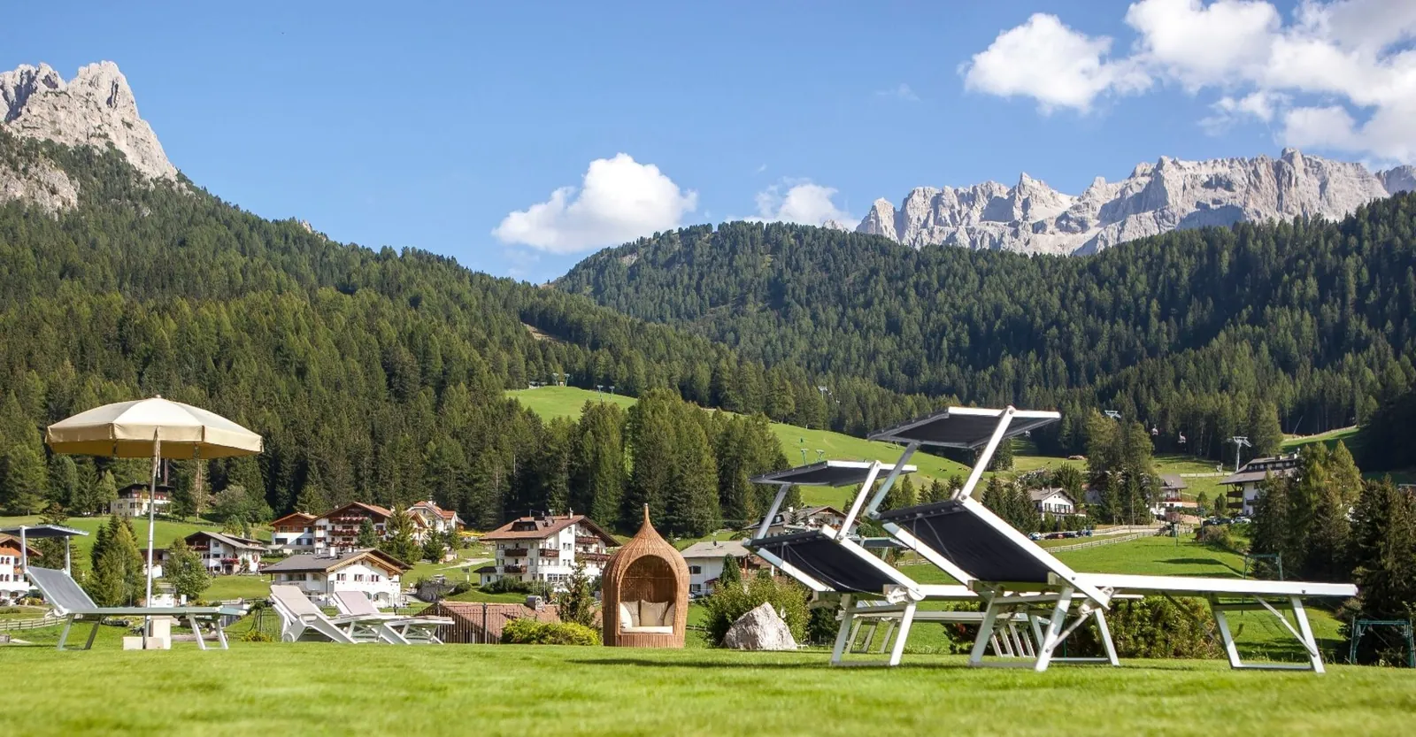 Liegewiese mit Sonnenliegen und Schirm, dahinter Wald und Dolomitengipfel